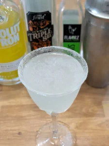 Lemon drop cocktail with a sugar rim in a stemmed glass, with citron vodka, triple sec and a cocktail shaker behind